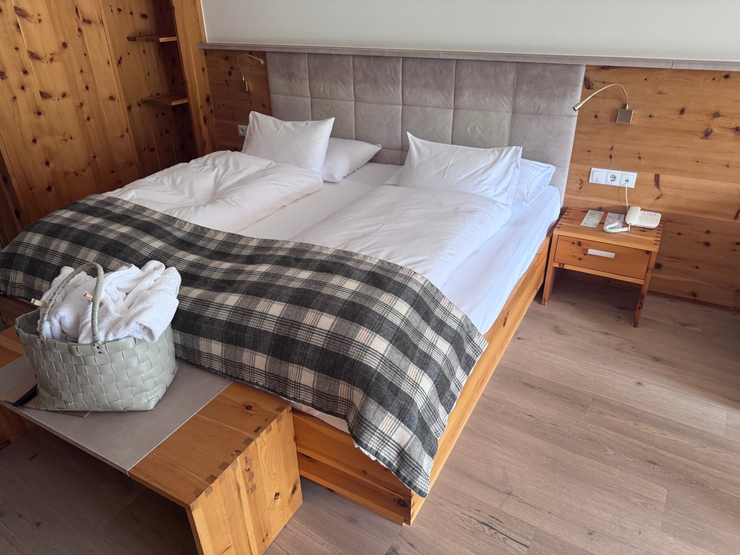 King-sized bed at Leitlhof Hotel with crisp white bedding, plush pillows, and a grey plaid throw blanket, with a woven basket containing rolled white towels placed on a wooden bench at the foot of the bed, set in a warm, wood-accented room.