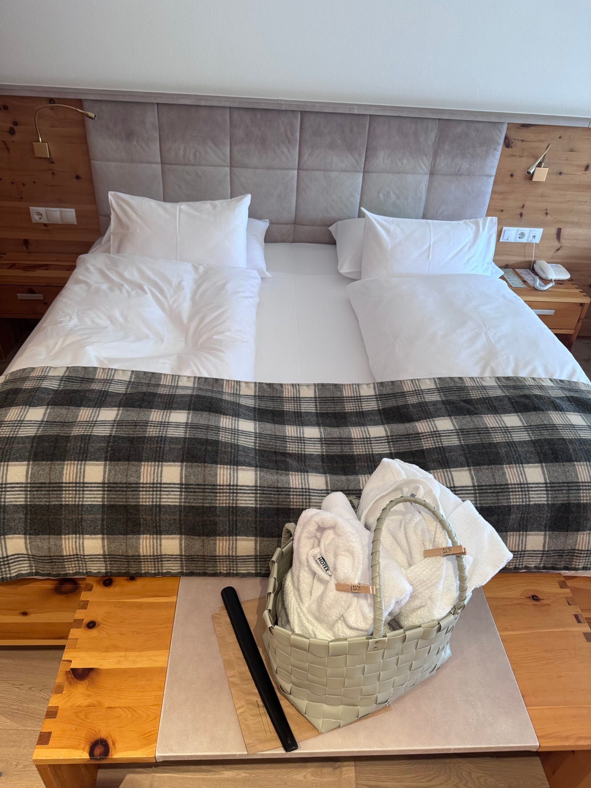 King-sized bed at Leitlhof Hotel with crisp white bedding, plush pillows, and a grey plaid throw blanket, with a woven basket containing rolled white towels placed on a wooden bench at the foot of the bed, set in a warm, wood-accented room.