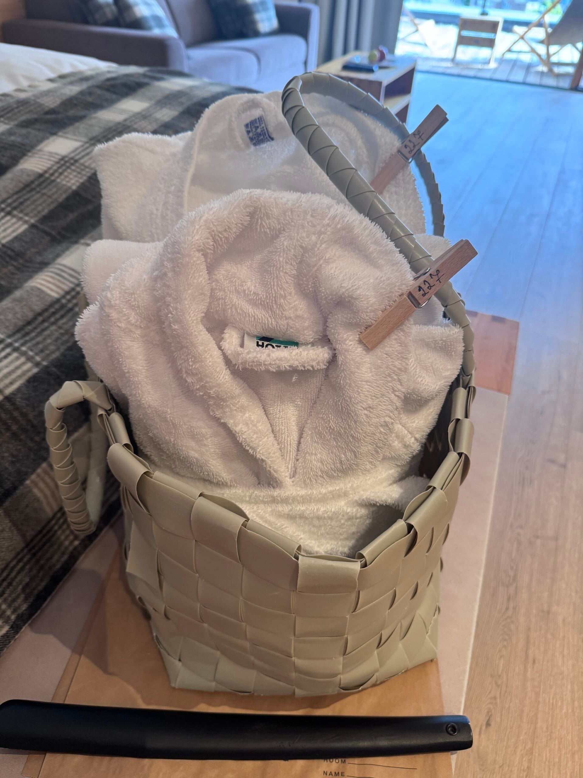 Woven beige basket containing neatly rolled white bath towels with ‘HOTEX’ label and wooden clothes pegs, placed on a wooden bench at the foot of the bed in Leitlhof Hotel’s cosy alpine-styled room.