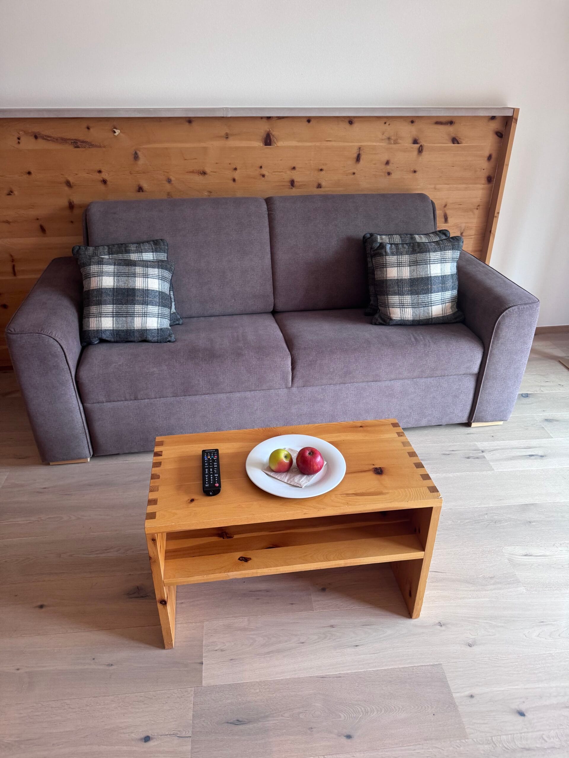 Grey cushioned sofa with plaid throw pillows in a Leitlhof Hotel room, set against wooden panel walls with a light wooden coffee table in front, holding a white plate with apples and a TV remote on a light wood floor.