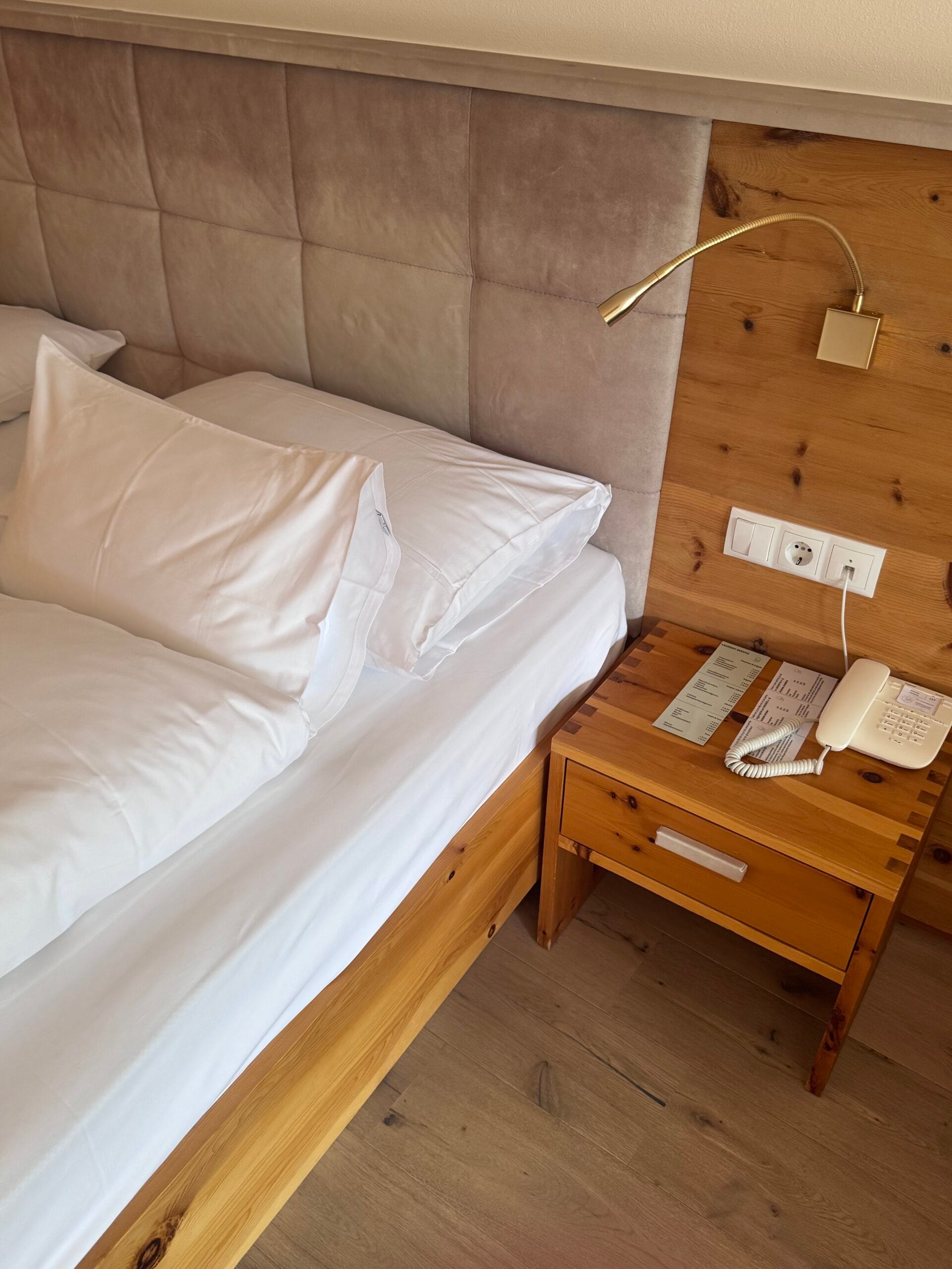 View of a comfortable bed with crisp white linens and plush pillows at Leitlhof Hotel, next to a rustic wooden bedside table holding a classic telephone and information cards, with a warm wood and beige upholstered headboard.