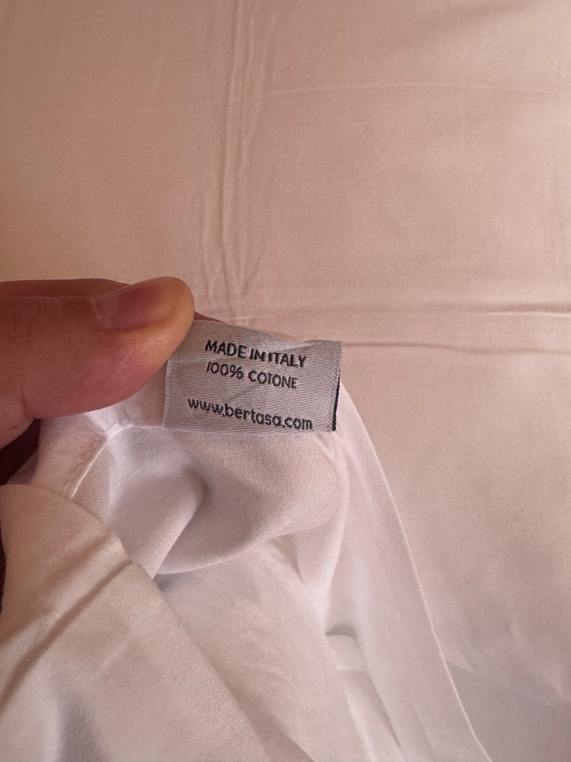 Close-up of a white fabric label reading ‘Made in Italy 100% Cotone’ from Bertasa.com, held in hand against the soft white cotton bedsheets at Leitlhof Hotel, showcasing the commitment to quality materials.