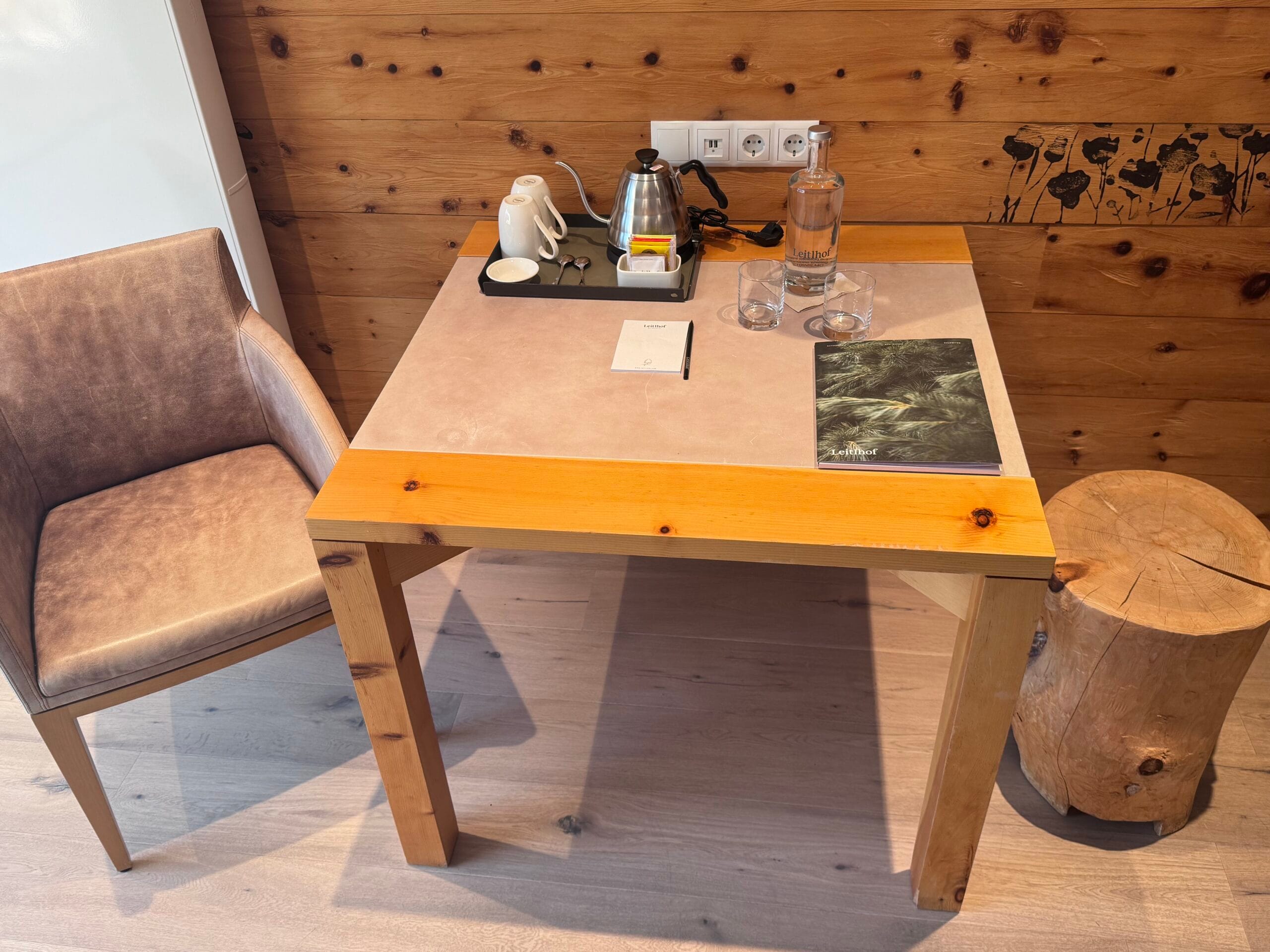 Small wooden desk in a Leitlhof Hotel room featuring a kettle, cups, bottled water, glasses, a notepad with a pen, and a Leitlhof-branded brochure, next to a beige cushioned chair and wood-panelled walls with floral art accents.