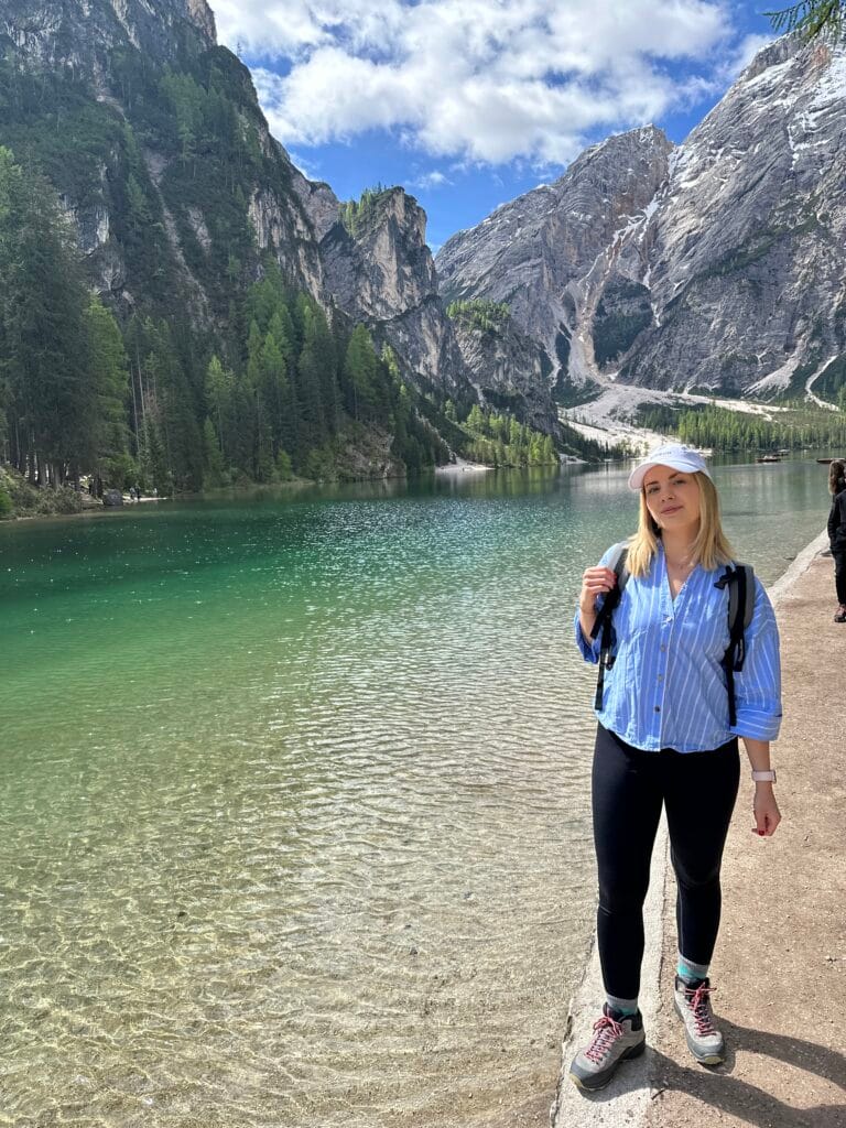 Woman standing beside the crystal-clear green waters of Lago di Braies, surrounded by forested mountains and rocky peaks in the Dolomites.