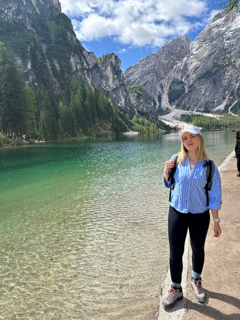 Woman standing beside the crystal-clear green waters of Lago di Braies, surrounded by forested mountains and rocky peaks in the Dolomites.