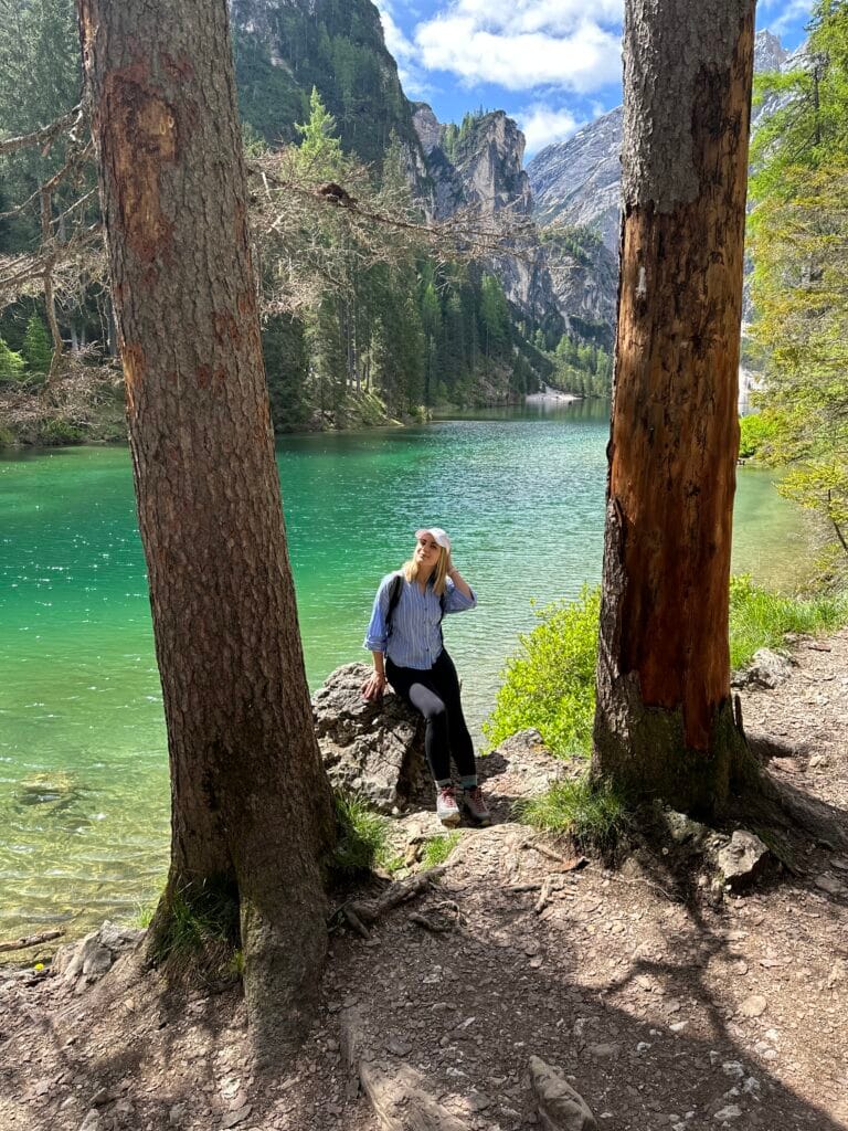 Woman sitting on a rock between two tall trees, overlooking the turquoise waters of Lago di Braies with alpine cliffs in the background.