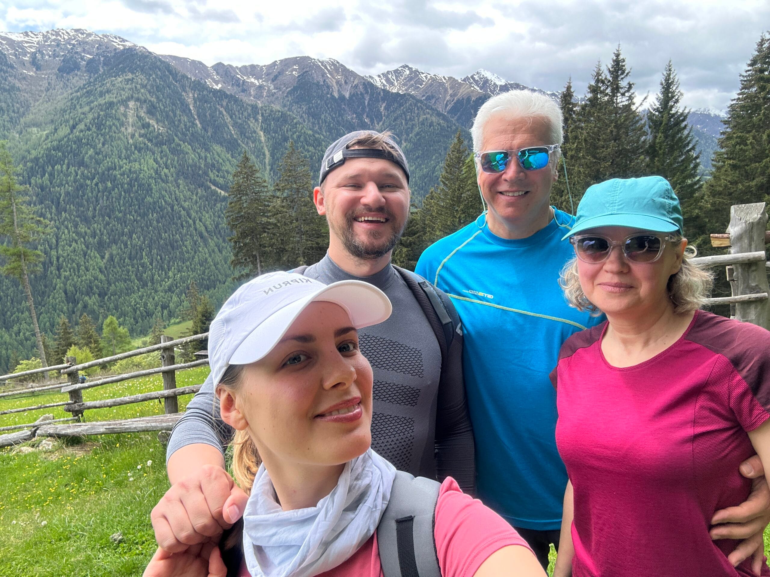 Four hikers smiling for a selfie with pine trees, grassy meadows, and rugged snow-capped mountains in the background in South Tyrol.