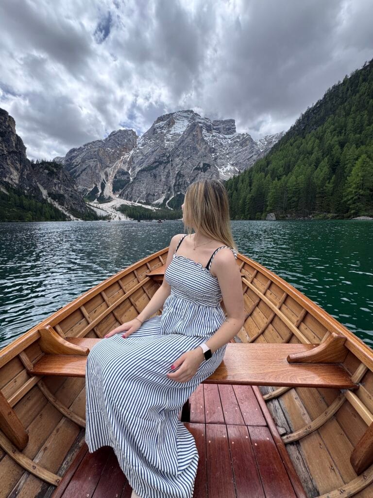Woman in a striped dress sitting in a wooden boat on Lago di Braies, surrounded by emerald water, pine forests, and towering Dolomite peaks.