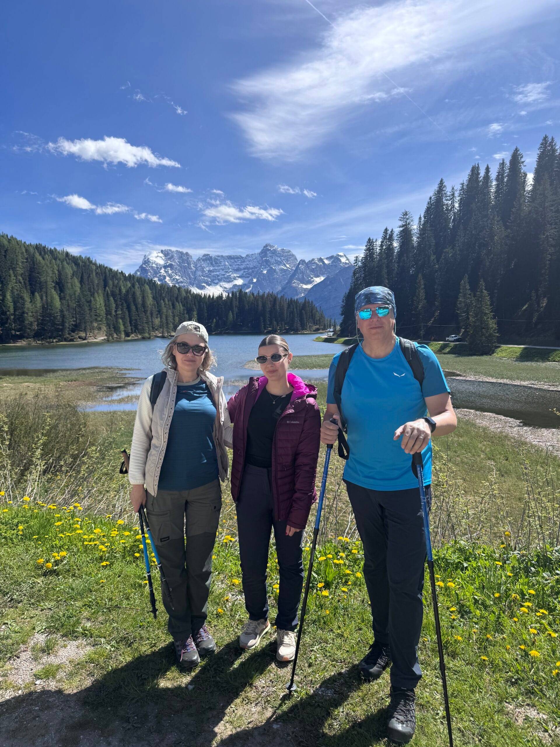 Three hikers posing in front of a lake with bright green grass, pine forests, and snow-covered Dolomite peaks under a blue sky.