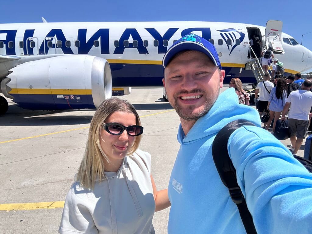 Smiling couple taking a selfie in front of a Ryanair airplane on the tarmac, ready to board under bright sunlight.