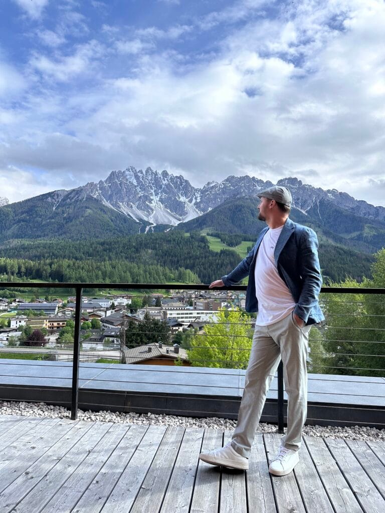 Man in a blazer and cap standing on a terrace overlooking a mountain village and the dramatic peaks of the Dolomites in South Tyrol.