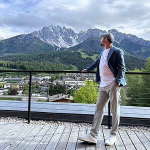 Man in a blazer and cap standing on a terrace overlooking a mountain village and the dramatic peaks of the Dolomites in South Tyrol.