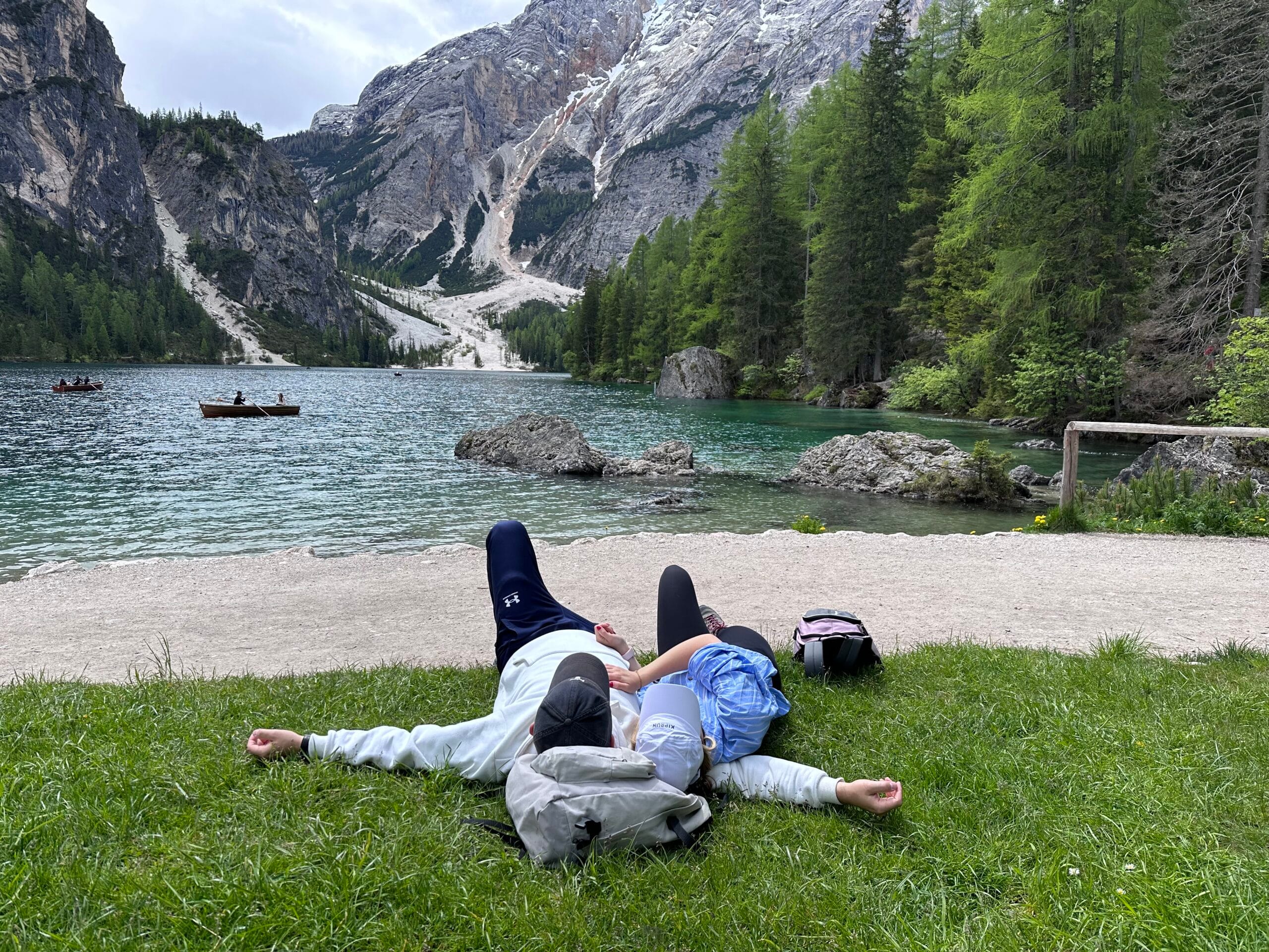 Two people lying on the grass by a mountain lake, relaxing with a view of water, trees, and rocky peaks.