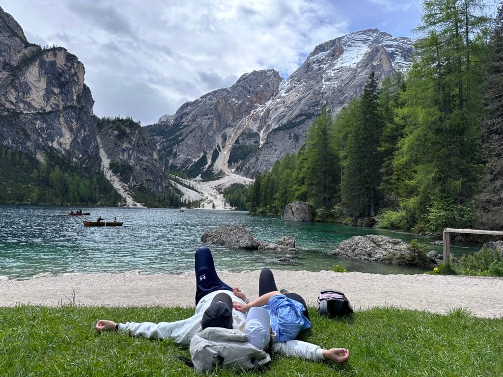 Two people lying on the grass beside Lago di Braies, backpacks nearby, gazing at the dramatic mountain peaks and pine forests in the Dolomites.