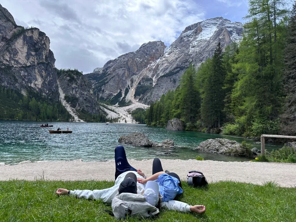Two people lying on the grass beside Lago di Braies, backpacks nearby, gazing at the dramatic mountain peaks and pine forests in the Dolomites.