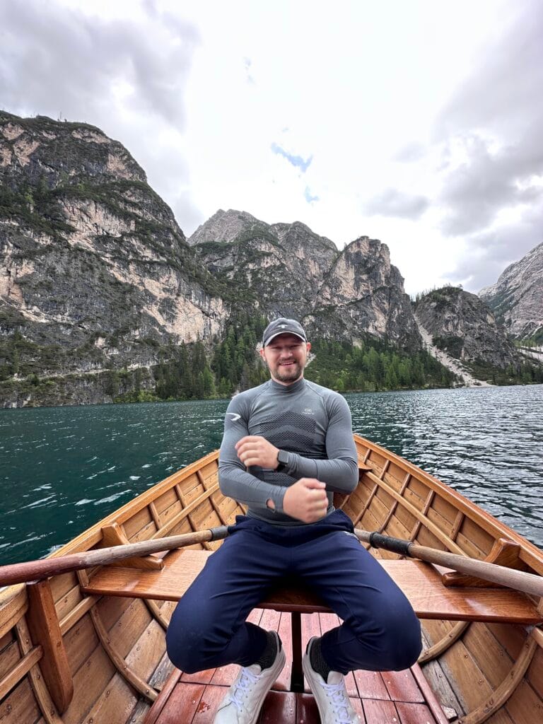 Man sitting at the front of a wooden rowboat on Lago di Braies, surrounded by emerald water and steep forested cliffs in the Dolomites.