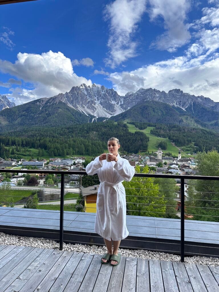 Woman in a white robe sipping coffee on a terrace with panoramic views of the Dolomite mountains and a charming Alpine village.
