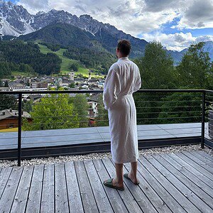 Woman in a white robe sipping coffee on a terrace with panoramic views of the Dolomite mountains and a charming Alpine village.