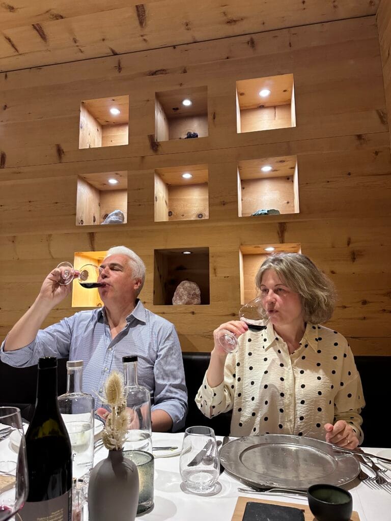 Mature couple enjoying red wine at a wooden-walled restaurant table, surrounded by glasses and decor, inside Leitlhof Hotel in South Tyrol.