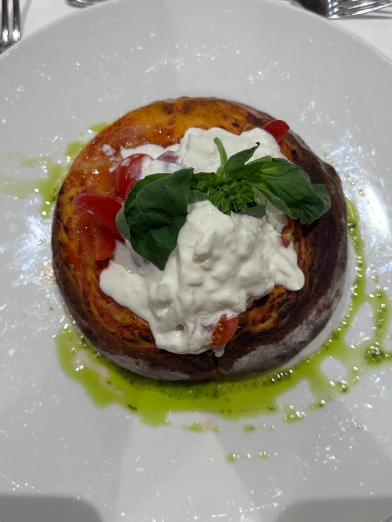 Fluffy tomato-based focaccia topped with burrata, cherry tomatoes, fresh basil, and a drizzle of green herb oil on a white plate.