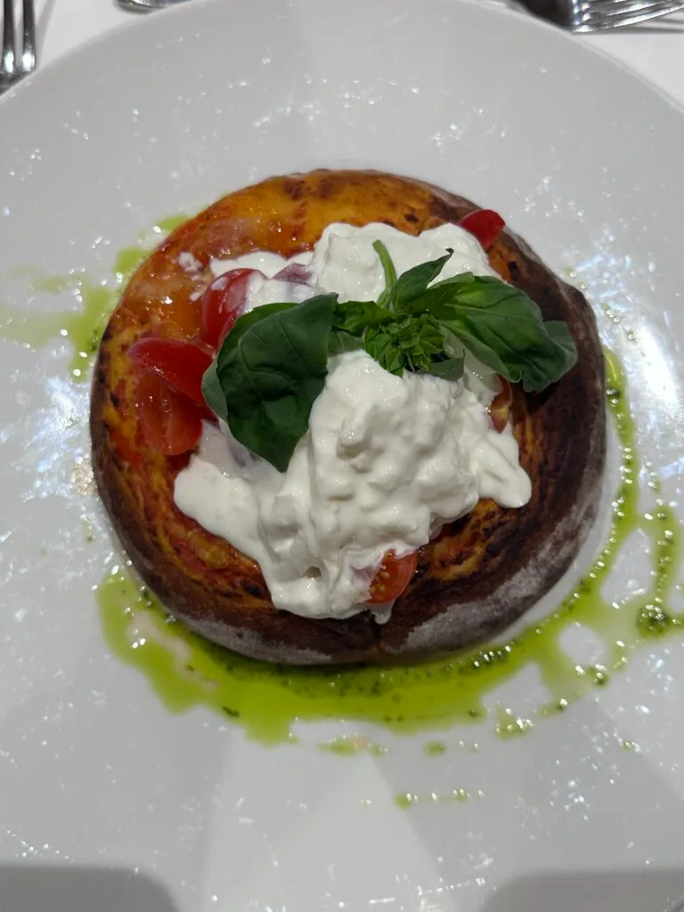 Fluffy tomato-based focaccia topped with burrata, cherry tomatoes, fresh basil, and a drizzle of green herb oil on a white plate.