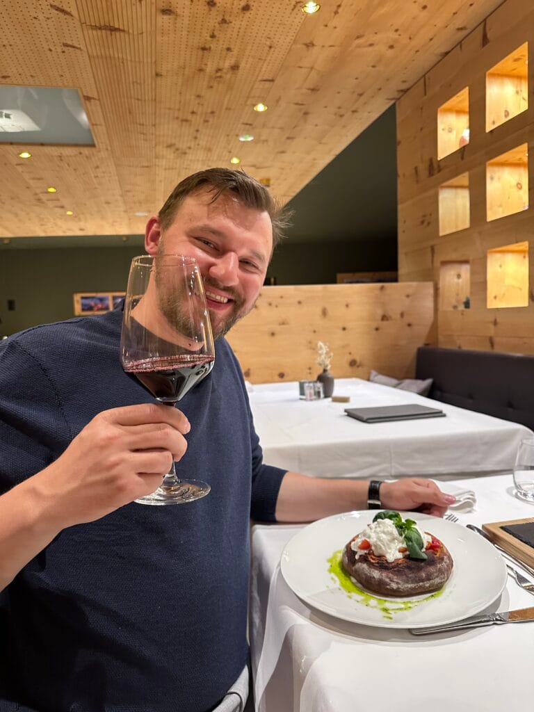 Man smiling while holding a glass of red wine at a wooden alpine restaurant table, with a burrata-topped focaccia dish in front of him.