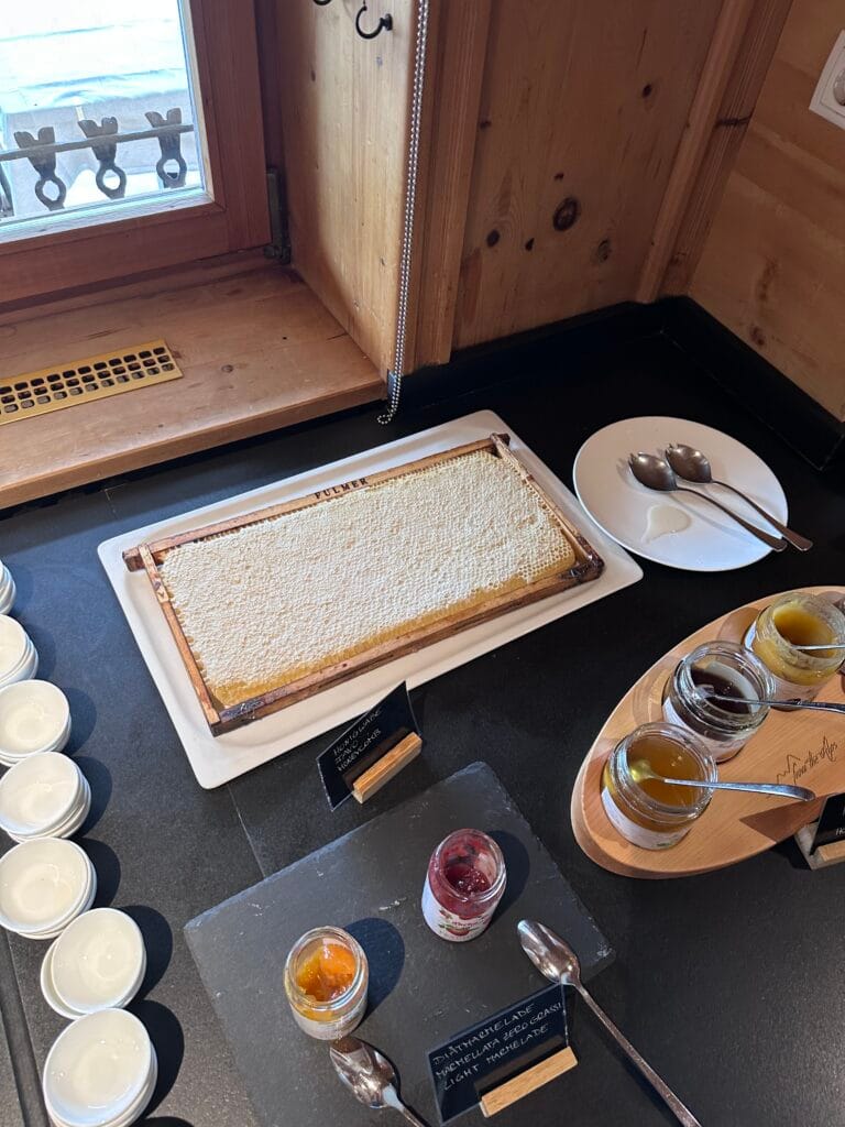 Fresh honeycomb served on a tray beside glass jars of honey and jam, with small plates and spoons arranged on a black breakfast buffet counter near a window.