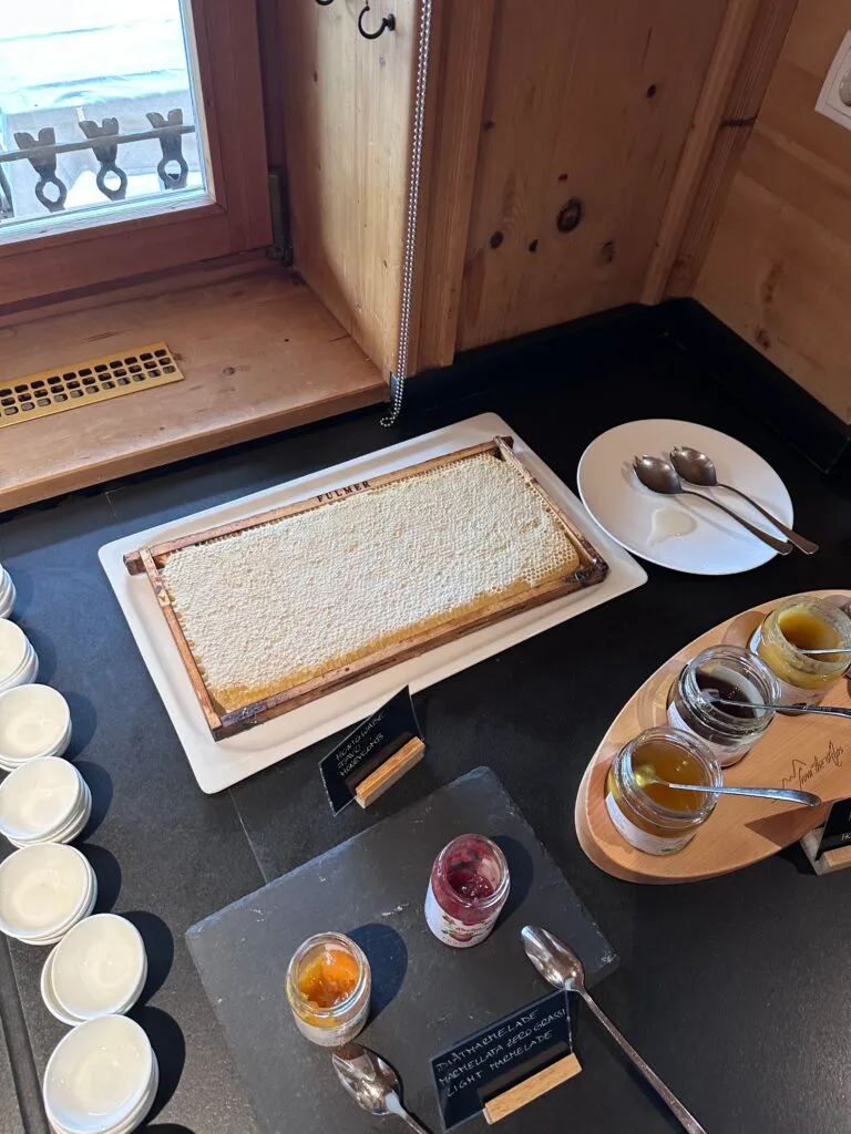 Fresh honeycomb served on a tray beside glass jars of honey and jam, with small plates and spoons arranged on a black breakfast buffet counter near a window.