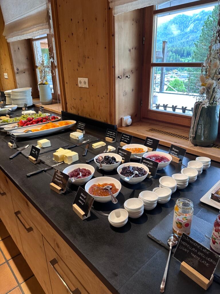 Breakfast buffet with assorted jams, fruit, butter, and cheeses arranged neatly on a black countertop, inside a wooden Alpine dining room with mountain views.