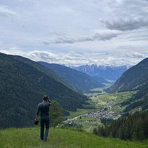 Man standing on a grassy ridge overlooking a vast alpine valley with forested slopes, winding roads, and distant snow-capped mountains in South Tyrol.