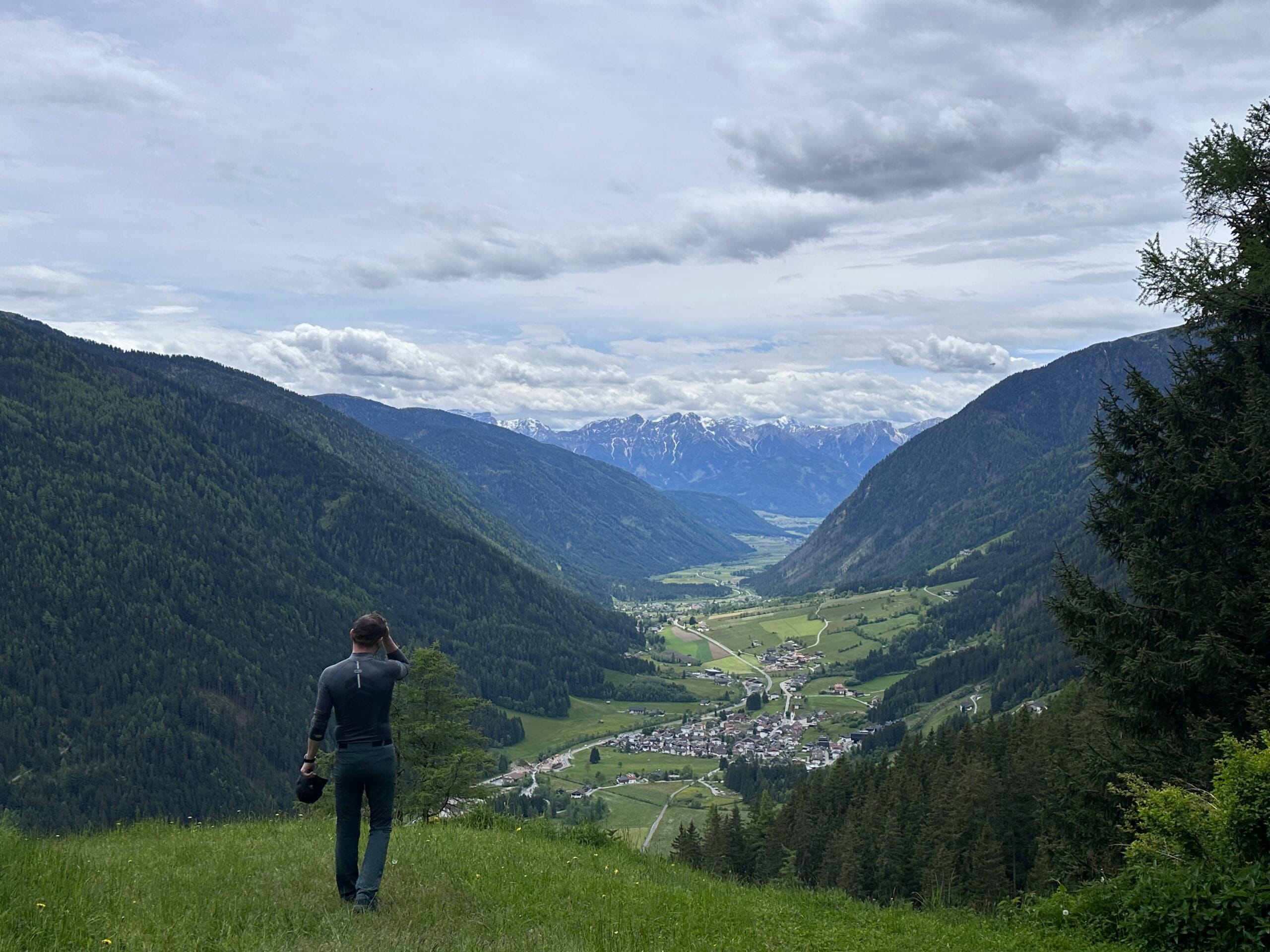 Man standing on a grassy ridge overlooking a vast alpine valley with forested slopes, winding roads, and distant snow-capped mountains in South Tyrol.