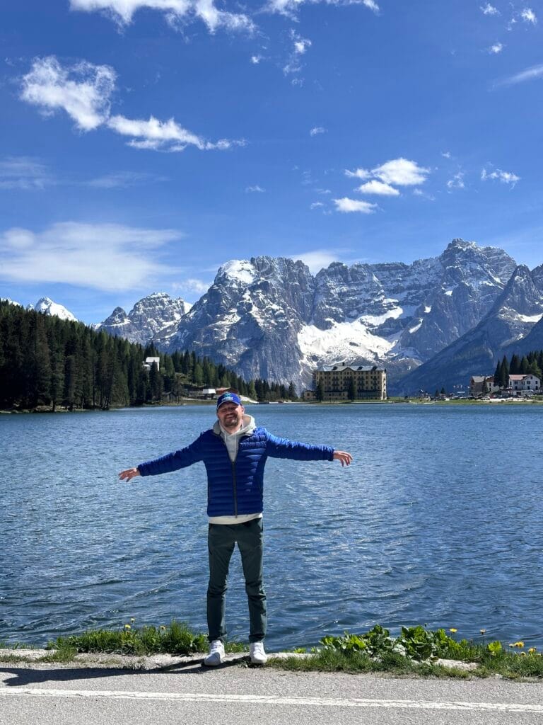 Man standing with arms outstretched by a mountain lake, smiling with snow-covered peaks and forested slopes in the background.