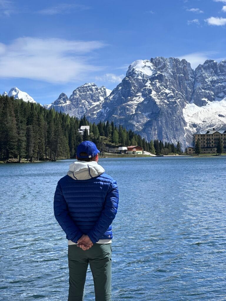 Man in a blue jacket and cap standing by a mountain lake, facing snow-capped peaks and forested slopes in the Italian Dolomites.