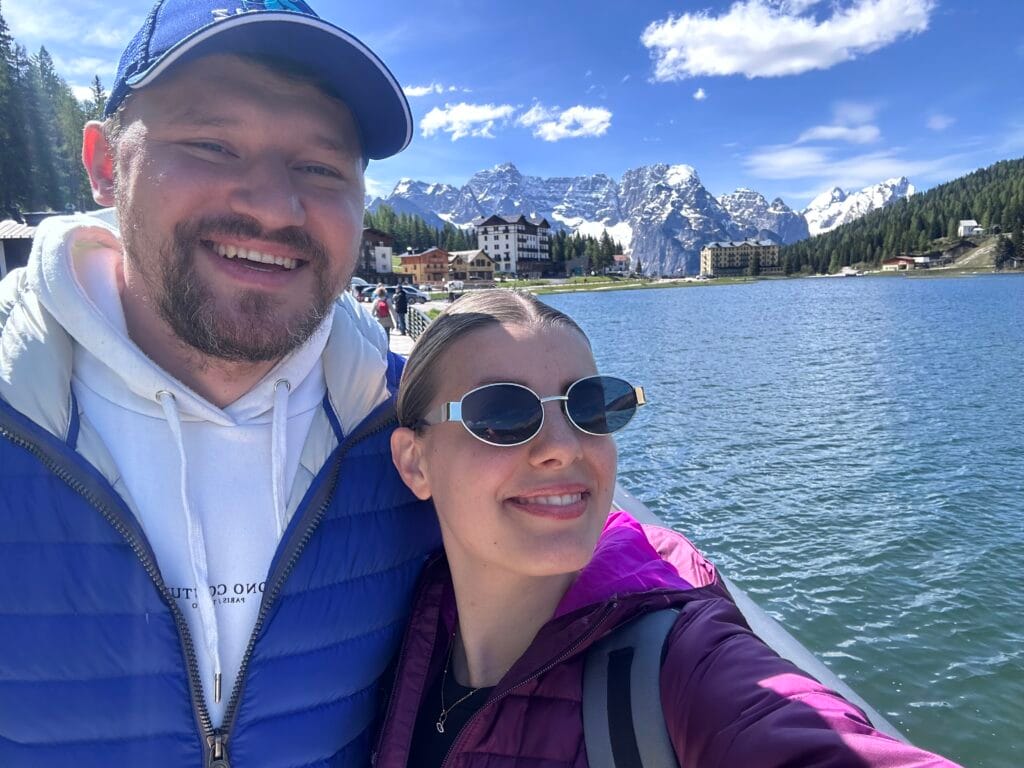 Smiling couple taking a selfie by Lake Misurina with alpine hotels, blue skies, and the snow-capped Dolomites in the background.