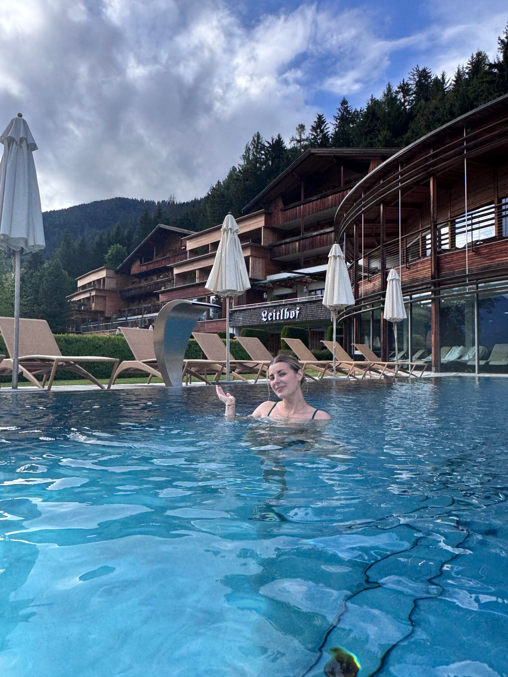 Woman relaxing in an outdoor heated pool at Leitlhof Hotel, surrounded by Alpine wooden architecture and mountain views in South Tyrol, Italy.