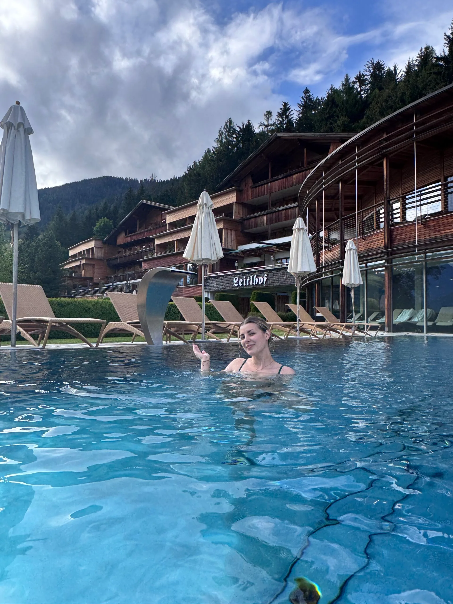 Woman relaxing in an outdoor heated pool at Leitlhof Hotel, surrounded by Alpine wooden architecture and mountain views in South Tyrol, Italy.