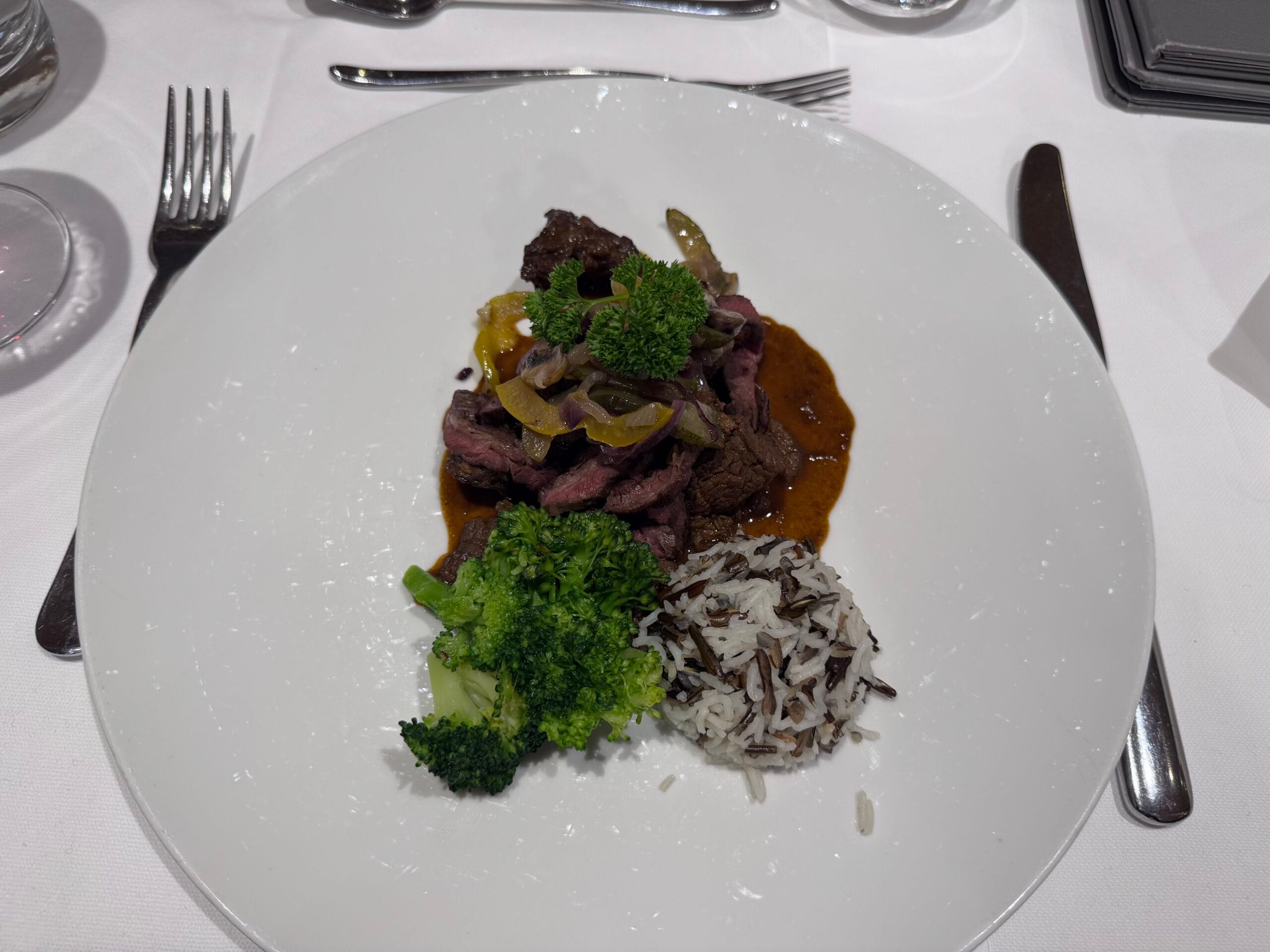 Plate of sliced steak with pepper sauce, steamed broccoli, and a side of wild rice, served on a white tablecloth with silver cutlery.