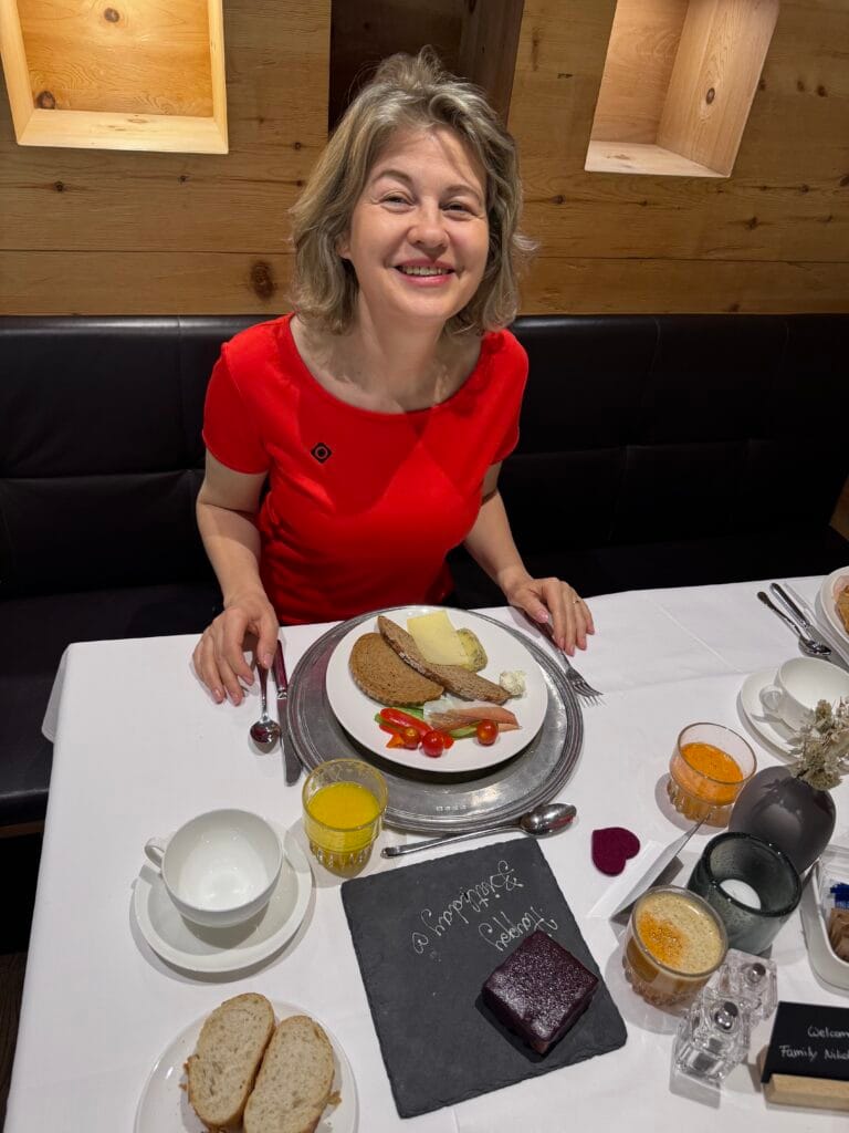 Woman smiling at a breakfast table with a plate of rye bread, cheese, smoked fish, cherry tomatoes, juice, and coffee inside a cosy wooden Alpine restaurant.