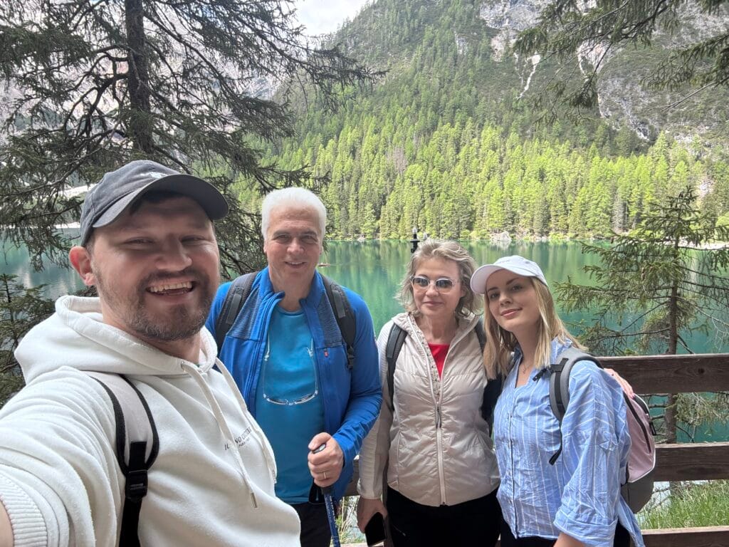 Group of four smiling hikers standing in front of the green waters of Lago di Braies, surrounded by pine-covered mountains in South Tyrol.