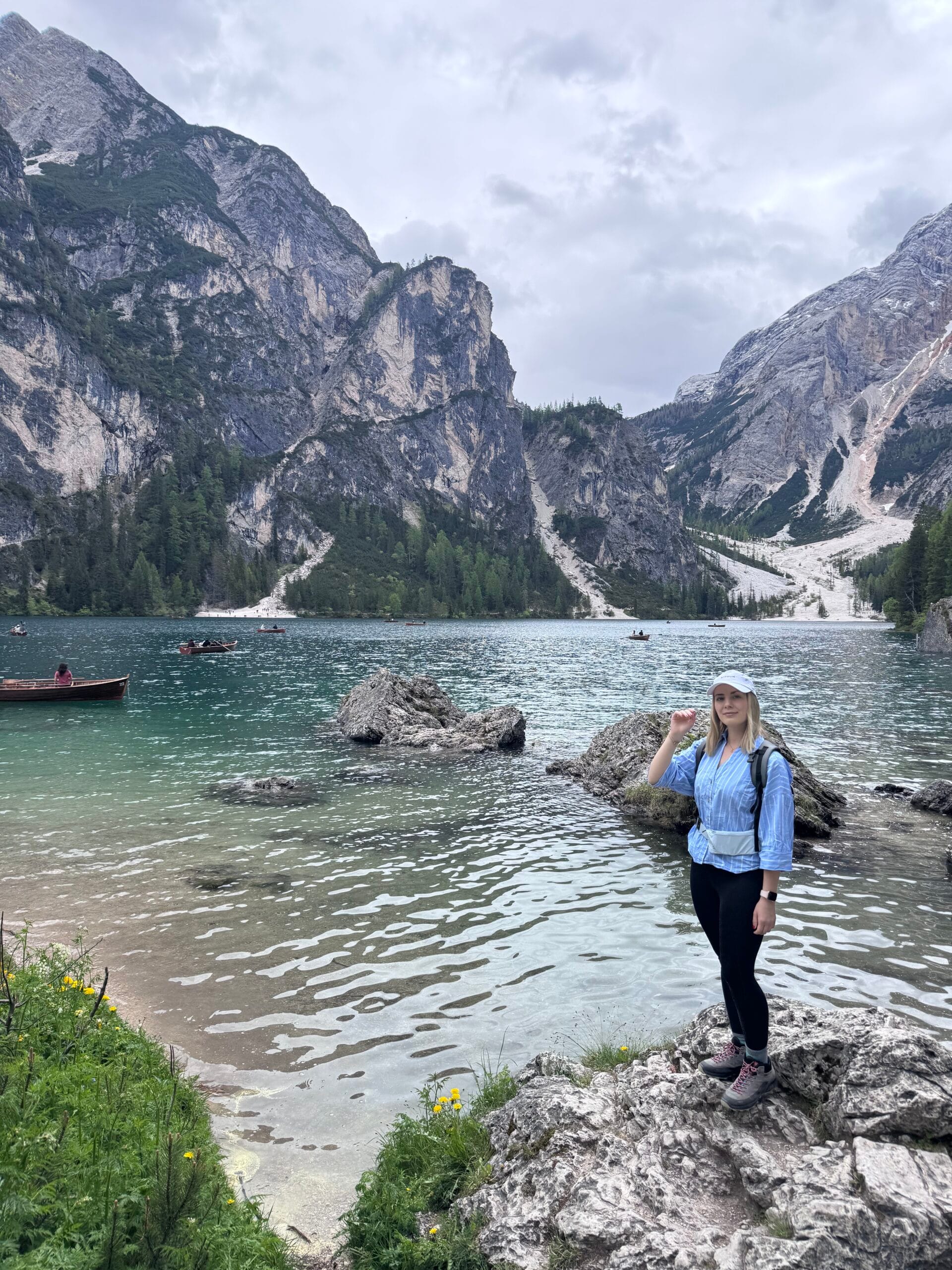 Woman standing on rocks at the edge of Lago di Braies, with turquoise water, rowboats, pine trees, and towering cliffs in the background.