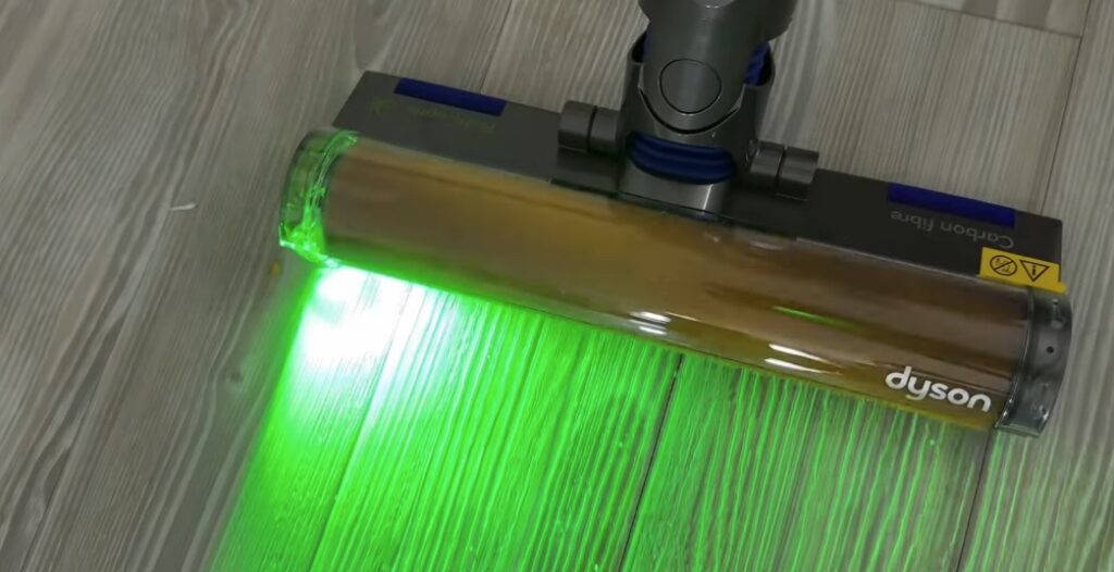 Close-up of a Dyson vacuum cleaner head with green laser light illuminating dust and debris on a light wood floor.