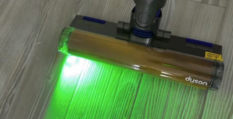 Close-up of a Dyson V12 vacuum cleaner head with green laser light illuminating dust and debris on a light wood floor.
