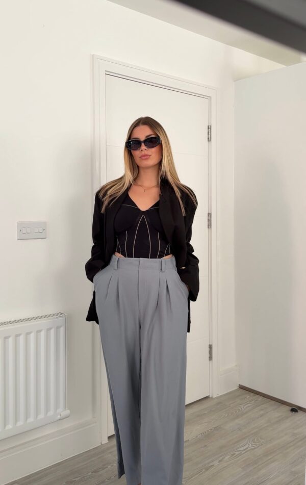 Female UGC creator wearing a black top and high-waisted grey trousers, posing indoors in a minimalist UK home setting.