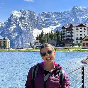 Young woman with sunglasses smiling by a lake with mountains in the background.