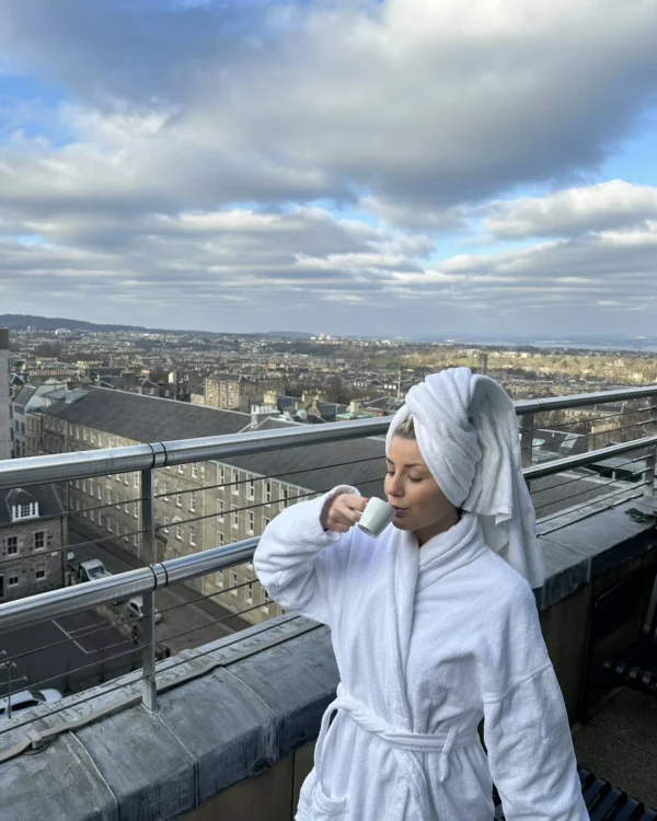 Woman in a white bathrobe drinking coffee on a balcony with panoramic city views in the background