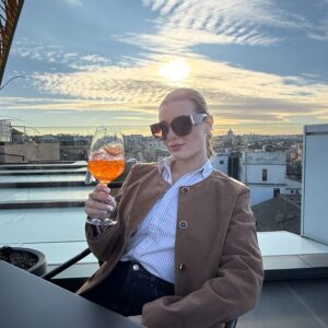 Woman with sunglasses holding a colourful cocktail on a rooftop at sunset.