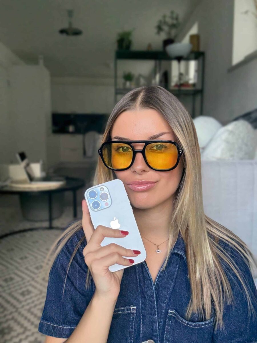 Velena Nikolova in a denim shirt and yellow sunglasses holding up her phone in a living room