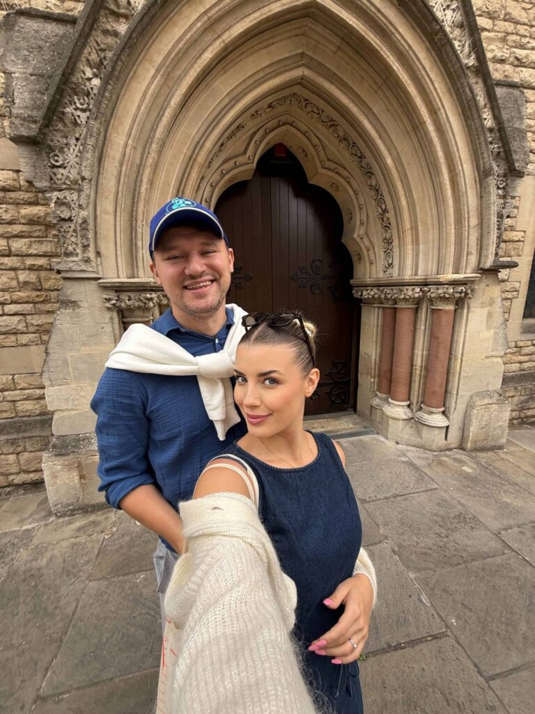 Dragos Nistor in a cap and Velena Nikolova in a denim top taking a selfie in front of a stone church doorway in Oxford