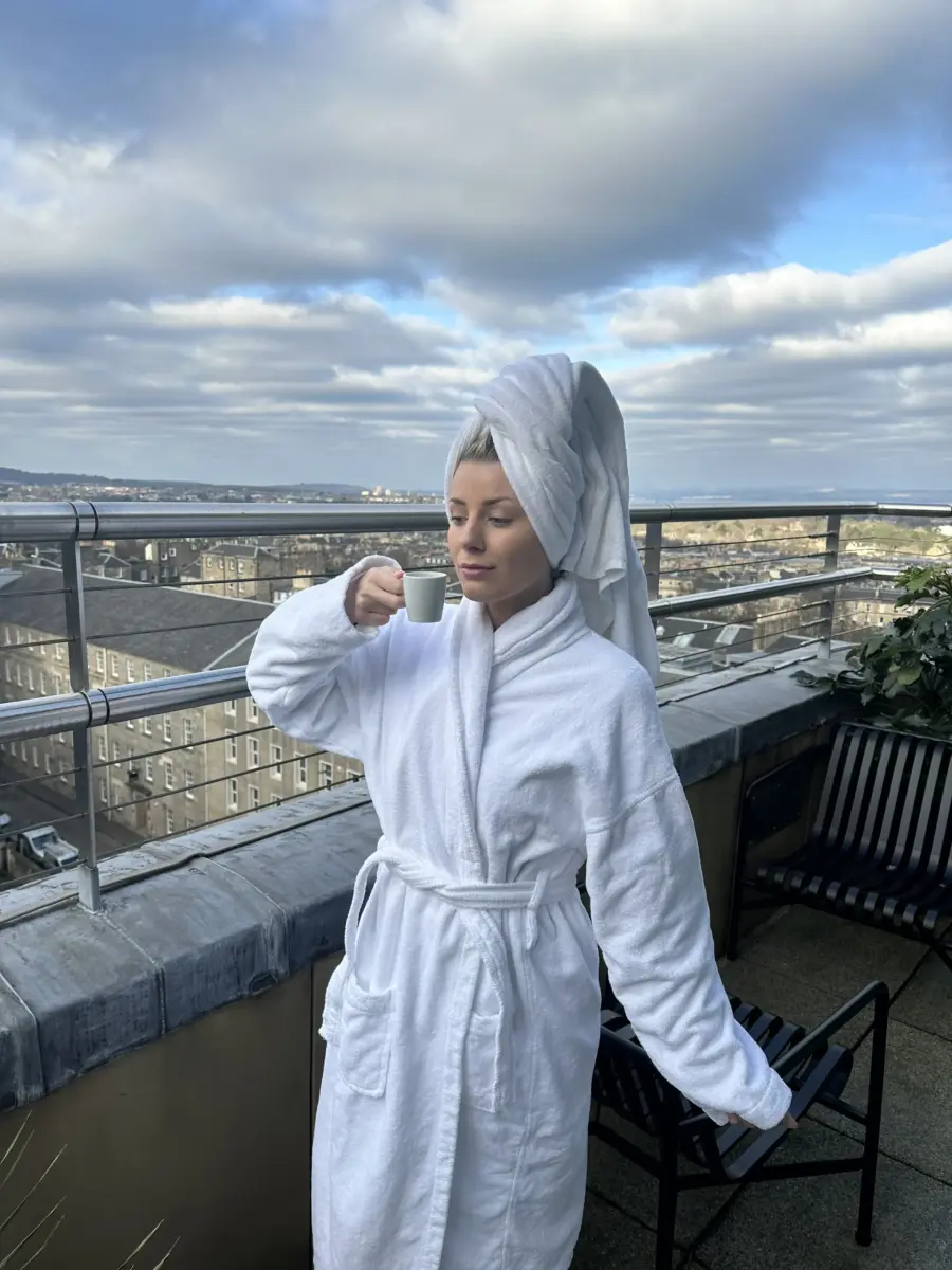 Velena Nikolova in a white bathrobe and a towel on her head drinking coffee on a hotel roof terrace above Edinburgh, wide