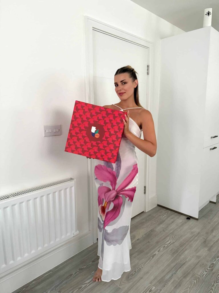 Velena Nikolova in a white dress with a pink flower print holding a red gift box in a white corridor
